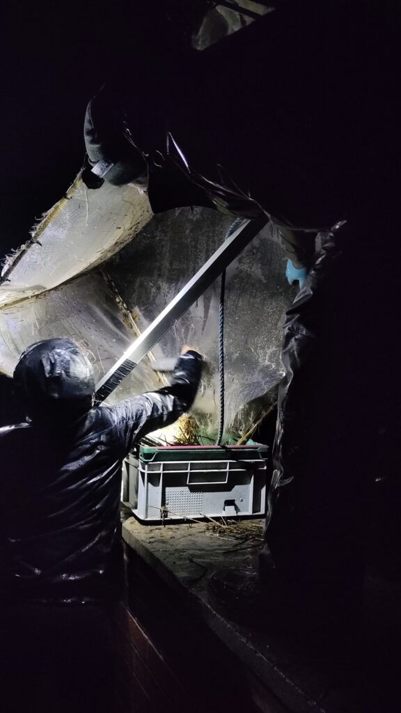 A person in a hooded waterproof jacket works at night on an eel-fishing setup, adjusting a large wet net structure above a plastic crate illuminated by a flashlight. The surrounding area is dark except for the focused light on the fishing gear